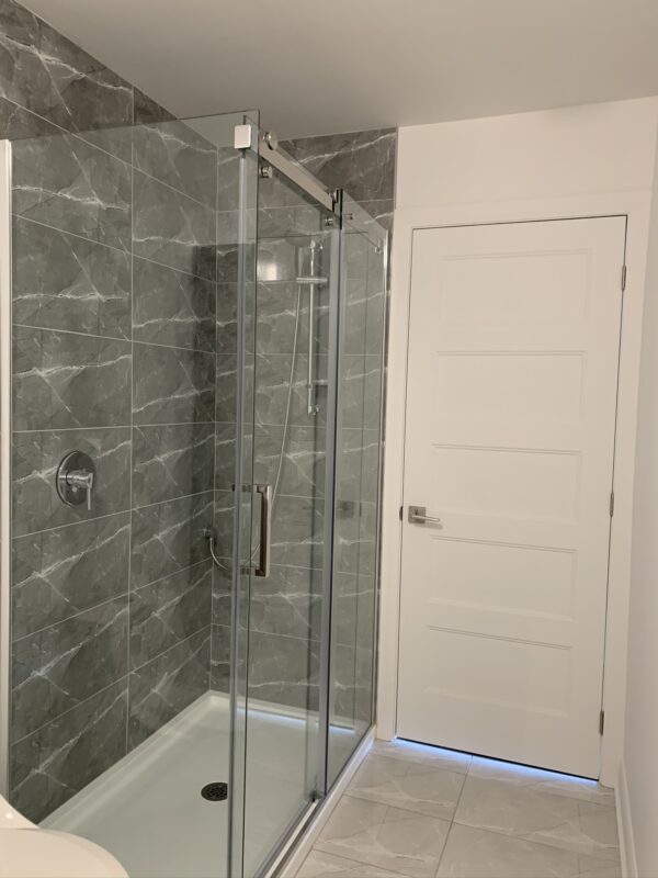 Salle de bain moderne avec douche en verre, carrelage gris et porte blanche dans un appartement à Mirabel, secteur St-Canut, disponible à partir du 1er juillet.