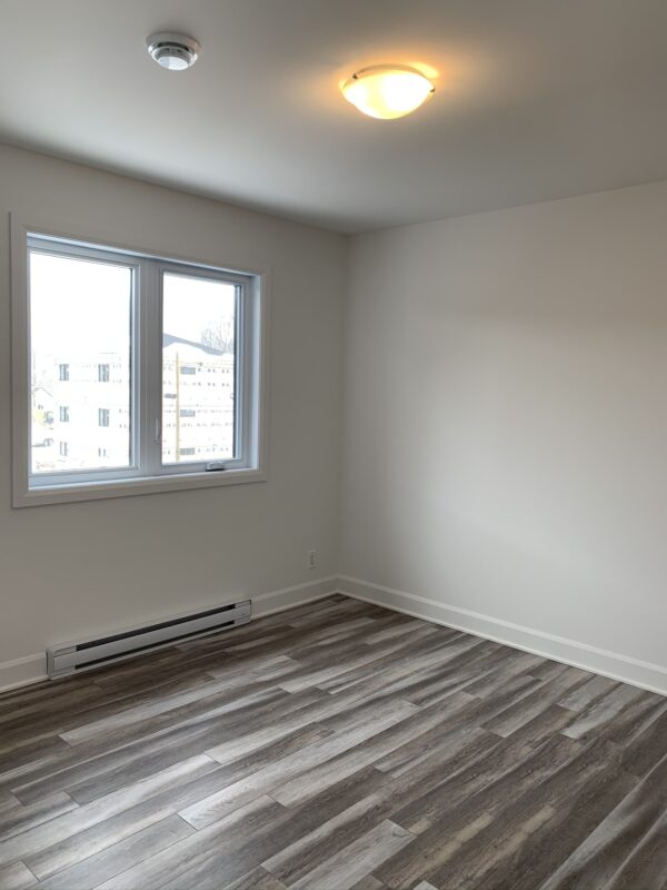 Pièce lumineuse avec deux fenêtres, sol en bois flottant, murs blancs, idéale pour un logement de 2 chambres à Mirabel, secteur St-Canut, disponible à partir du 1er juillet.