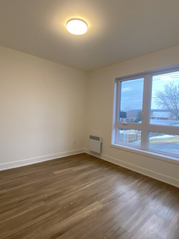 Appareil photo dans une pièce vide avec mur blanc, grande fenêtre et sol en bois clair. Idéal pour la gestion immobilière à Montréal, cette propriété offre un espace accueillant et moderne pour locataires ou investisseurs.