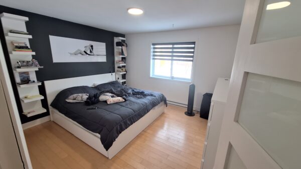 Chambre lumineuse à louer dans un appartement à Saint-Calixte, avec lit double, étagères et fenêtres offrant une vue agréable, idéal pour un séjour confortable.