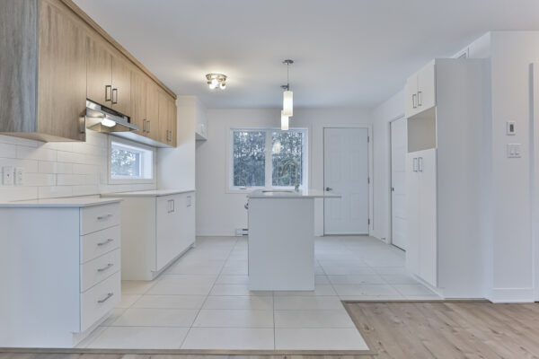 Cuisine moderne et lumineuse avec comptoir central, armoires en bois clair, plancher en bois et fenêtres offrant une vue sur l'extérieur, idéale pour location à Repentigny.