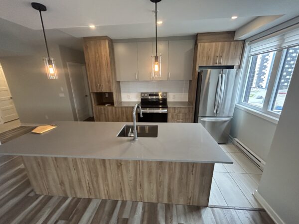 Appareil photo capturant une cuisine contemporaine avec îlot central, armoires en bois clair, électroménagers modernes, et grande fenêtre laissant entrer la lumière naturelle.