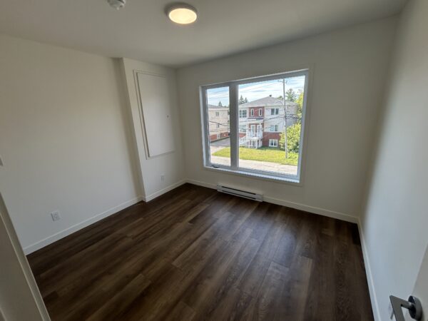 Chambre lumineuse avec grande fenêtre offrant vue sur le quartier. Parquet en bois, murs blancs, espace idéal pour un appartement à Saint-Jérôme.