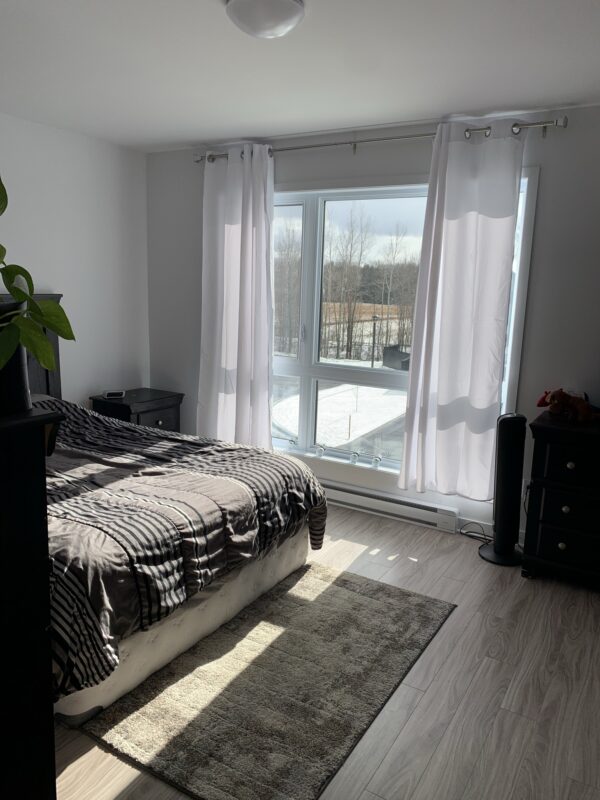 Chambre à coucher moderne avec grande fenêtre offrant une luminosité naturelle abondante, idéale pour un appartement à Mirabel, situé rue Siméon-Lamarche, en juillet.