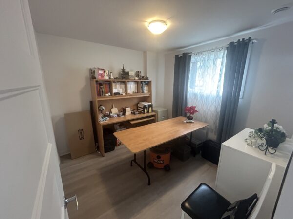 Intérieur d'un appartement d'une chambre à Lachute, avec un bureau, une étagère, et une fenêtre laissant entrer la lumière naturelle.