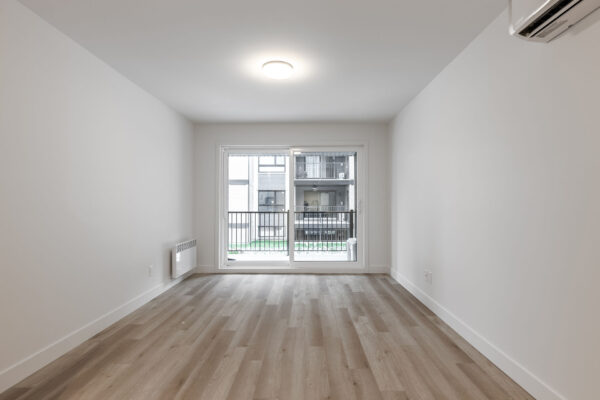 Vue d'un salon lumineux avec grande fenêtre donnant sur un balcon, dans un appartement de deux chambres à Saint-Jérôme, disponible en juillet.