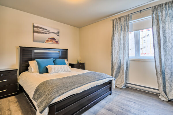 Chambre lumineuse avec lit double, oreillers bleus et décor moderne, dans un logement à louer à Mirabel, idéal pour étudiants ou jeunes professionnels.