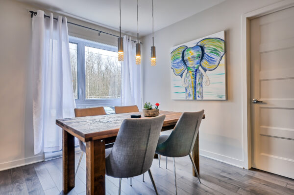Salle à manger lumineuse avec table en bois, chaises grises, grande fenêtre et décoration moderne, idéal pour un logement à Mirabel.